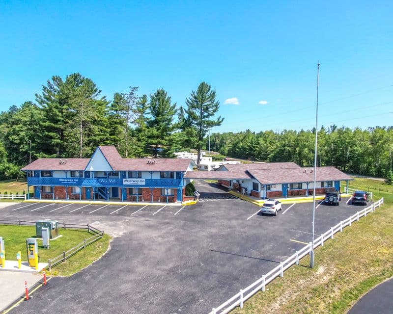 Aerial view of the Waterway Inn — a two-storey blue and white motel with balcony walkways and signage over the entrance, set behind a large parking lot ringed by pines.